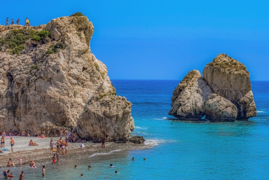 Beachgoers near rugged rocks at Aphrodite’s Rock under a blue sky.